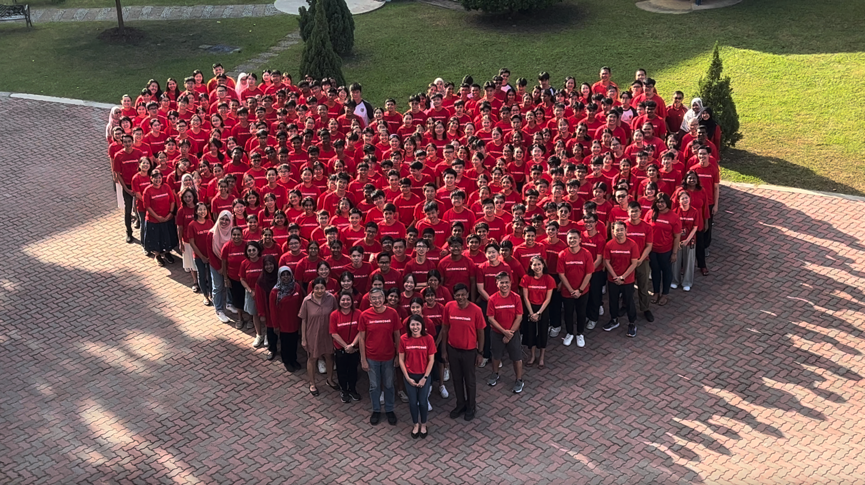 Large group of people wearing red shirts arranged in a heart shape on a brick courtyard, viewed from above.